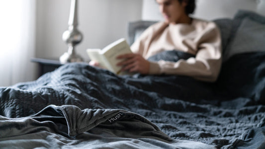 A man sitting in bed covered in grey Hush Classic Weighted Blanket while reading a book.