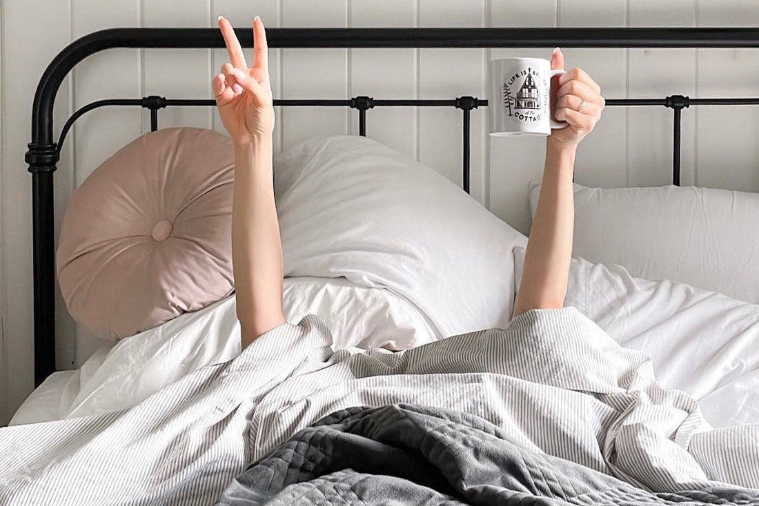A person laying in bed holding up a coffee mug in one hand and making a 'Peace Sign' with the other.