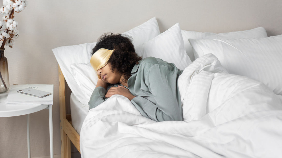 A woman of color sleeps soundly on a white bed while wearing a gold-colored silk sleep mask.