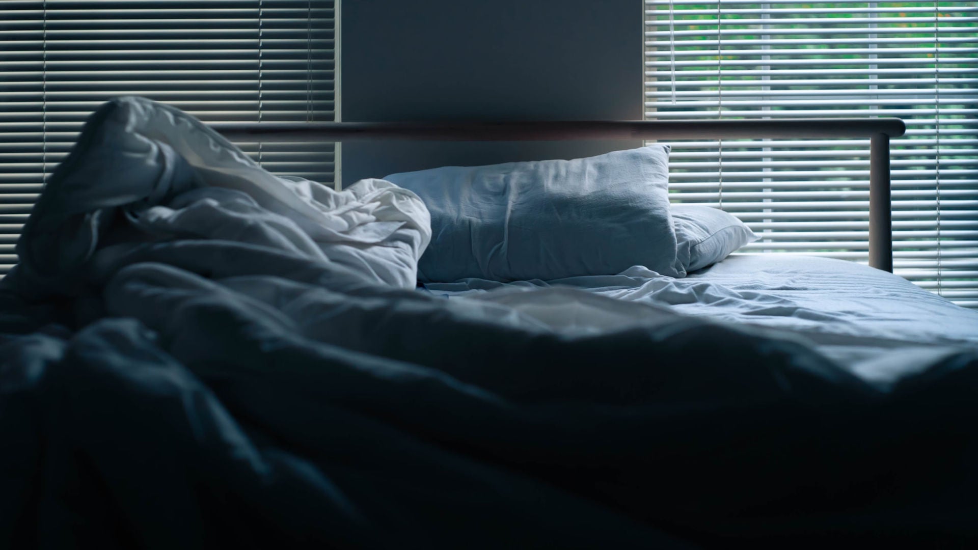 A bed with grey beddings with two windows with blinds behind.