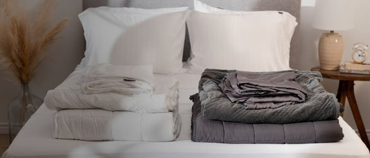 Two sets of Hush Classic weighted blankets with covers and pillowcases in white and grey colors neatly folded and stacked by color at the end of the bed.
