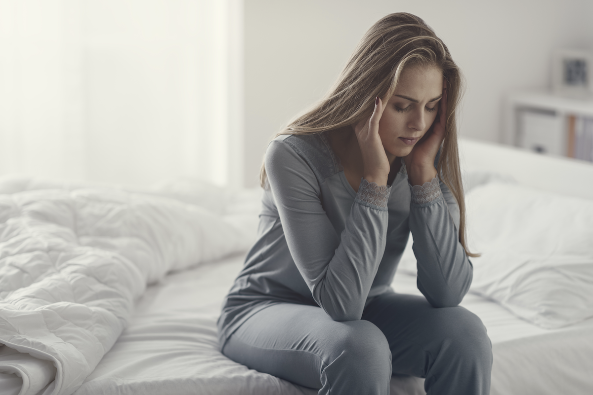 waking up with anxiety: Woman sitting on a bed with her eyes closed while rubbing her temples